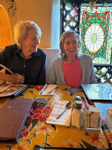 Two older women smiling together at a restaurant lunch table, enjoying a shared moment of companionship and connection.